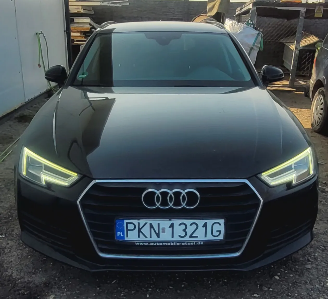 Czarny samochód marki Audi z przodu, z widocznym logo czterech pierścieni na atrapie chłodnicy. Tablica rejestracyjna z białym tłem i czarnym napisem PKN 1321G, po lewej stronie niebieski pasek z oznaczeniem PL oraz symbolem Unii Europejskiej. Poniżej tablicy adres strony www.automobile-atael.de. Samochód stoi na podwórku z żwirową nawierzchnią, w tle biały budynek z zielonym wężem, stos desek i kamienna ściana. Po prawej stronie częściowo widoczny inny pojazd.