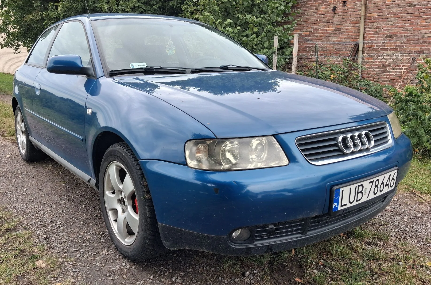 Niebieski samochód marki Audi zaparkowany na żwirowym podłożu przed ceglanym murem i zielonymi krzewami. Widoczny jest przód i lewy bok pojazdu. Samochód posiada polską tablicę rejestracyjną o numerze LUB 78646.