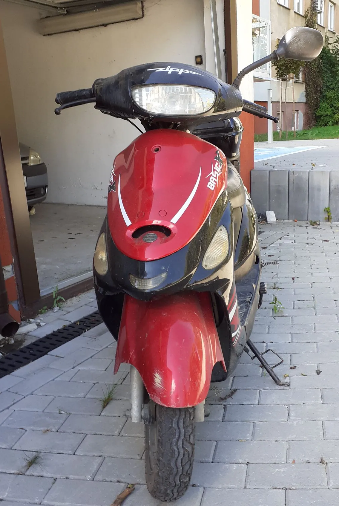 Czerwono-czarny skuter marki Zipp, model Basic, zaparkowany na kostce brukowej przed budynkiem. Pojazd jest ustawiony przodem do obiektywu, widoczny jest przedni reflektor, kierownica oraz przednie koło.