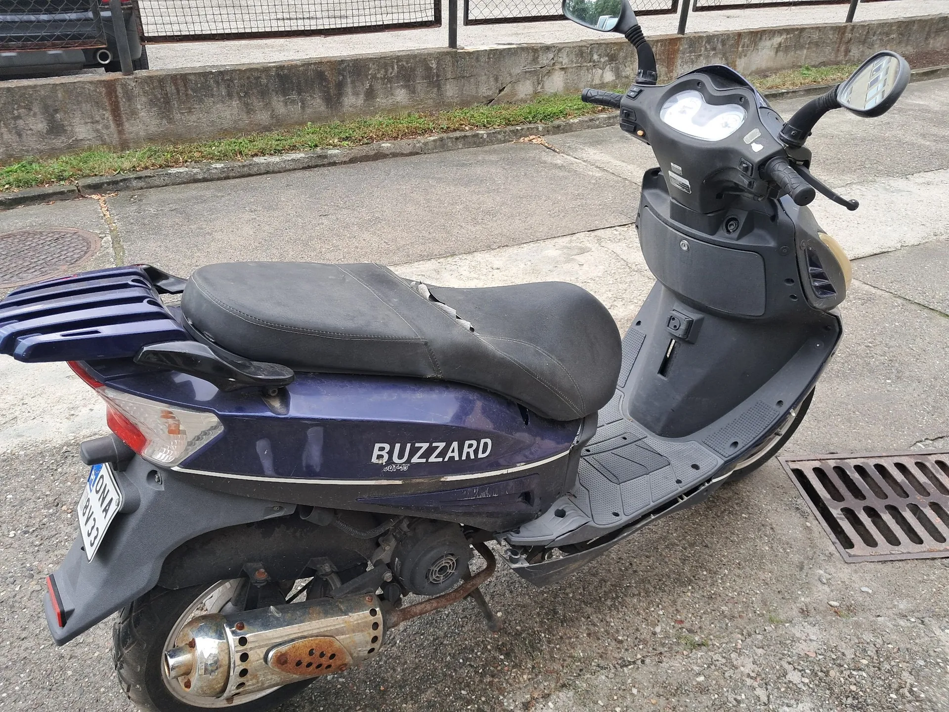 Granatowy skuter marki Buzzard zaparkowany na betonowym podłożu. Pojazd jest widoczny z profilu od tyłu, posiada czarne siedzenie, metalowy bagażnik z tyłu oraz chromowany wydech. Na boku skutera widnieje biały napis „BUZZARD”. W tle znajduje się krawężnik, trawnik oraz metalowy płot.