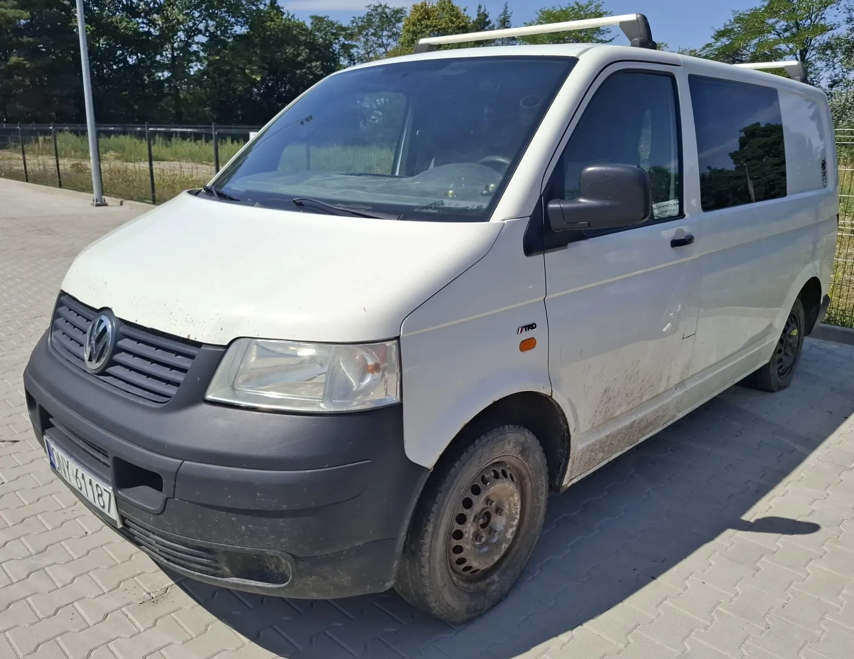 Biały samochód dostawczy marki Volkswagen Transporter zaparkowany na kostce brukowej. Pojazd posiada czarny zderzak przedni, czarne lusterka boczne oraz bagażnik dachowy. Na przednim zderzaku widoczna jest tablica rejestracyjna o numerze ANY 61187. Karoseria w dolnej części drzwi pasażera jest zabrudzona błotem.