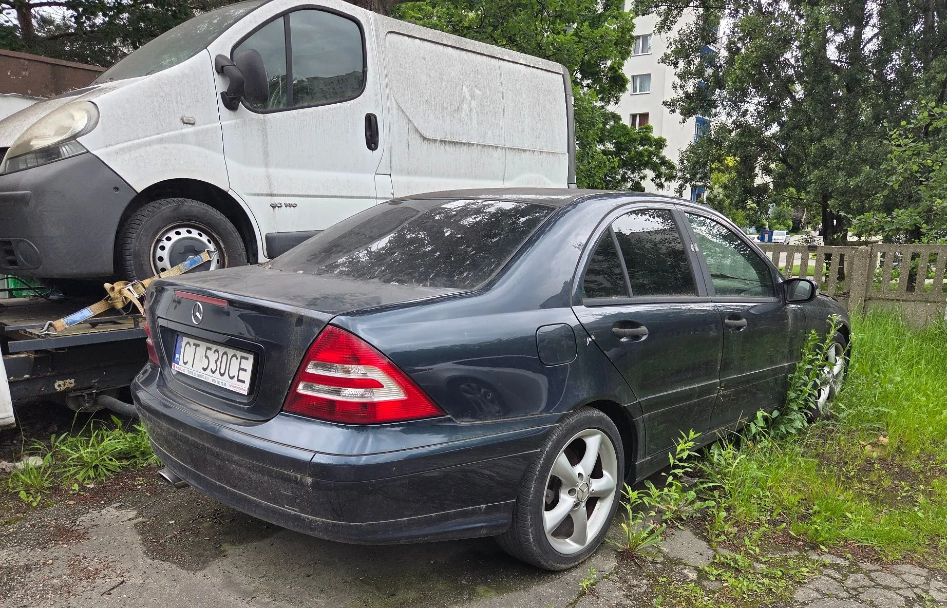 Zdjęcie przedstawiające ciemnogranatowy samochód osobowy marki Mercedes-Benz zaparkowany na trawiastym poboczu obok białego vana. Widoczny jest tył samochodu z tablicą rejestracyjną o numerze CT 530CE. Pojazd jest zakurzony, a wokół niego rośnie wysoka trawa i chwasty. W tle widoczne są drzewa oraz fragment wielopiętrowego budynku mieszkalnego.