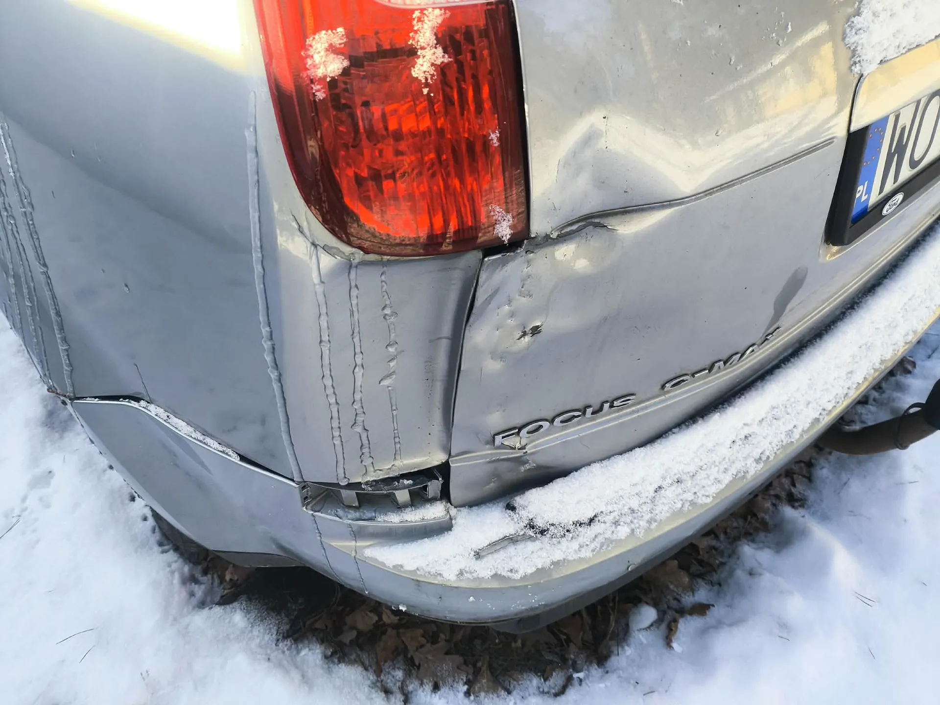 Zbliżenie na zniszczony tył srebrnego samochodu marki Ford Focus C-MAX. Widoczne są wgniecenia i zadrapania na błotniku oraz zderzaku. Czerwone światło tylnego reflektora częściowo pokryte śniegiem. Na prawo od reflektora tablica rejestracyjna z oznaczeniem PL i literami WO. Na zderzaku widoczny napis FOCUS C-MAX. Samochód stoi na śnieżnej powierzchni z widocznymi liśćmi na ziemi.