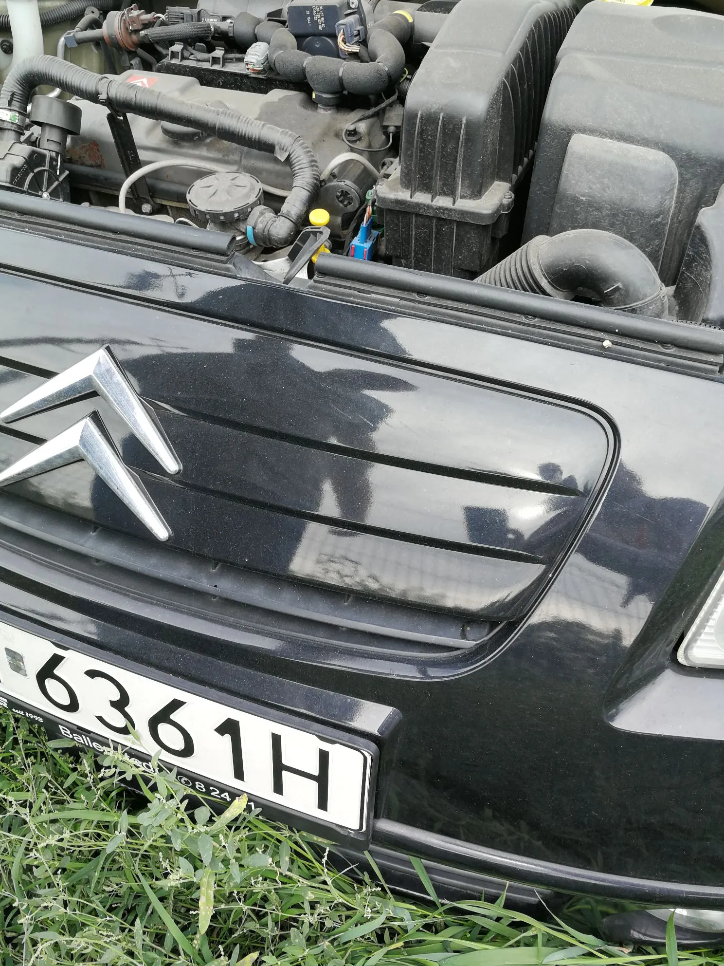 Zbliżenie na przód czarnego samochodu marki Citroën. Widoczne jest srebrne logo marki na masce oraz fragment tablicy rejestracyjnej z numerem 6361H. Nad maską znajduje się otwarta komora silnika z widocznymi elementami mechanicznymi, przewodami i obudową filtra powietrza. Samochód stoi na trawie.