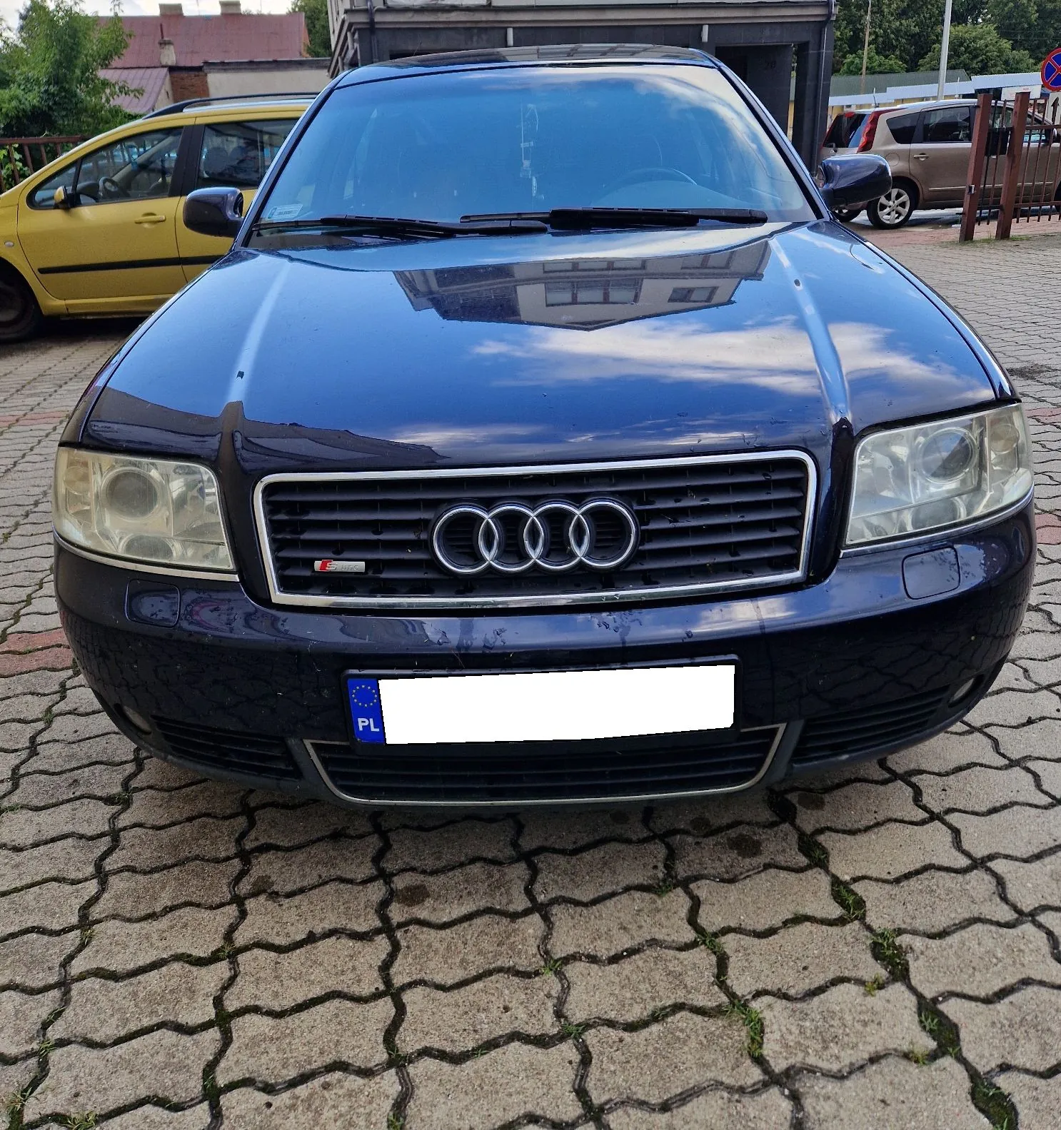 Zdjęcie przodu ciemnoniebieskiego samochodu marki Audi zaparkowanego na kostce brukowej. Samochód ma srebrne logo czterech pierścieni na grillu oraz białą tablicę rejestracyjną z oznaczeniem PL. W tle widoczny jest żółty samochód oraz brązowy samochód typu kombi.
