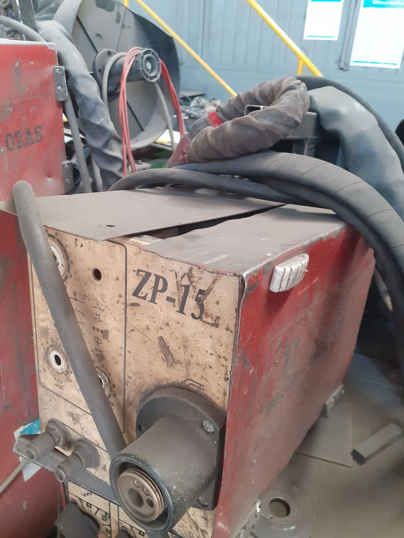 Zbliżenie na fragment czerwonej maszyny przemysłowej. Na metalowej, lekko zabrudzonej powierzchni widnieje czarny napis 'ZP-15'. Wokół urządzenia znajdują się grube, czarne kable oraz przewody. W tle widoczne są elementy konstrukcyjne i fragmenty innych urządzeń.