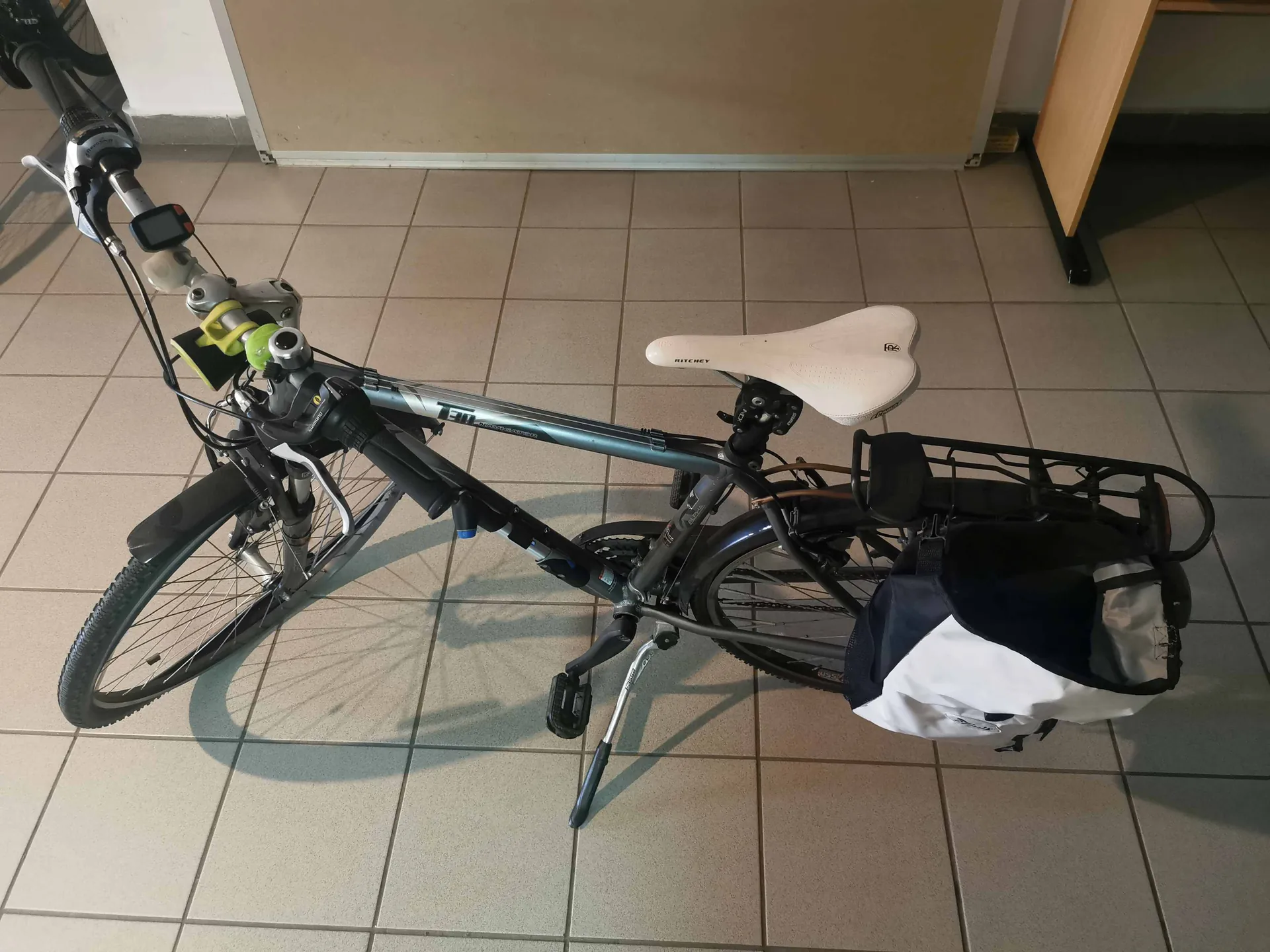 Srebrny rower miejski z białym siodełkiem, zaparkowany na jasnej, kafelkowej podłodze. Rower jest wyposażony w błotniki, koszyk z przodu z zamontowanym urządzeniem elektronicznym, a z tyłu posiada czarny bagażnik z przymocowaną biało-czarną torbą transportową. Rower stoi na stopce.