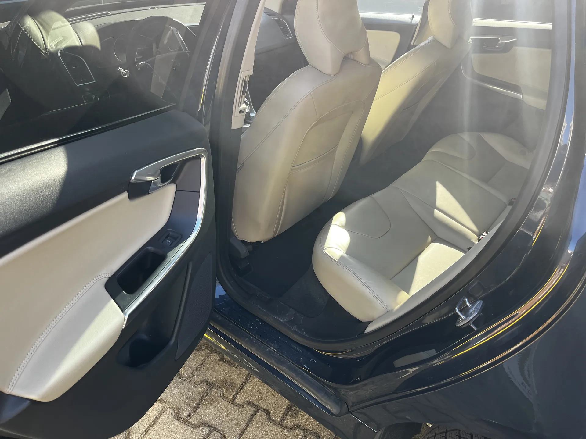 Wnętrze samochodu z otwartą przodem pasażerską drzwiami. Widoczne są jasnobrązowe skórzane siedzenia, czarno-beżowa obudowa drzwi z uchwytem i otworem na napoje. W tle kierownica i deska rozdzielcza. Samochód stoi na brukowanej powierzchni, słońce rzuca cienie na wnętrze. Brak czytelnego tekstu.