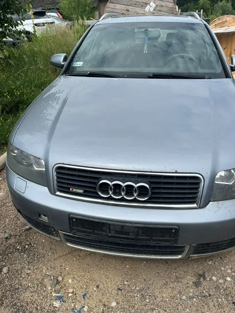 Srebrny samochód marki Audi widziany od przodu, zaparkowany na żwirowym podłożu. Widoczny jest charakterystyczny grill z czterema pierścieniami logo Audi oraz mały emblemat S-line. W tle widać zieleń oraz fragmenty innych pojazdów i konstrukcji drewnianych.
