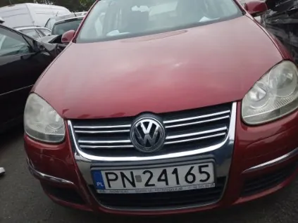 Czerwony samochód marki Volkswagen z przodu, z widocznym logo na atrapie chłodnicy. Tablica rejestracyjna z białym tłem i czarnym napisem 'PN 24165' oraz oznaczeniem 'PL' po lewej stronie. W tle częściowo widoczne inne zaparkowane pojazdy.