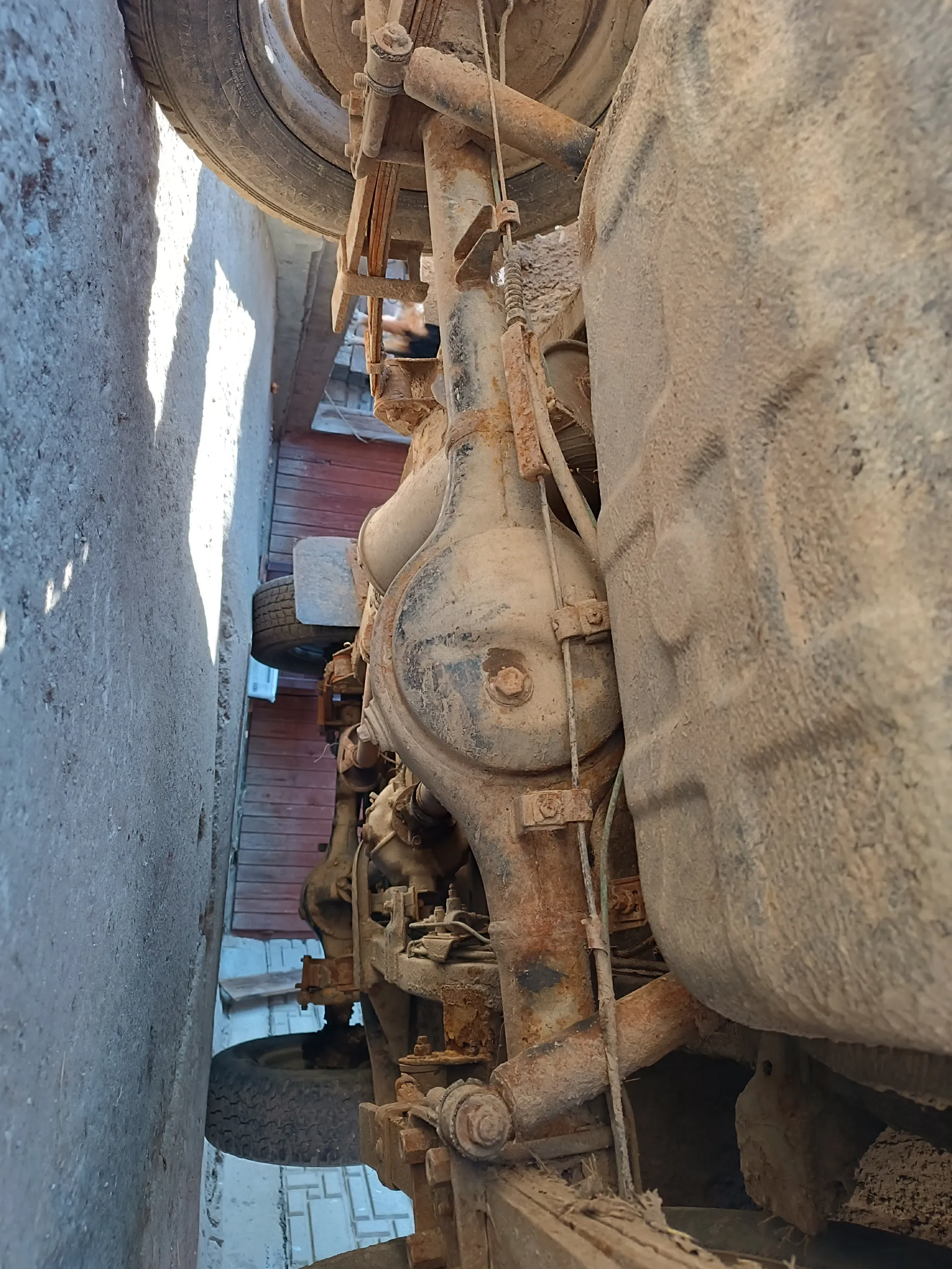 Podwozie pojazdu z brudnym zbiornikiem paliwa, rdzewnymi elementami metalowymi, zawieszeniem i oponami. W tle betonowa podłoga, czerwone drewniane drzwi i ceglana ściana.