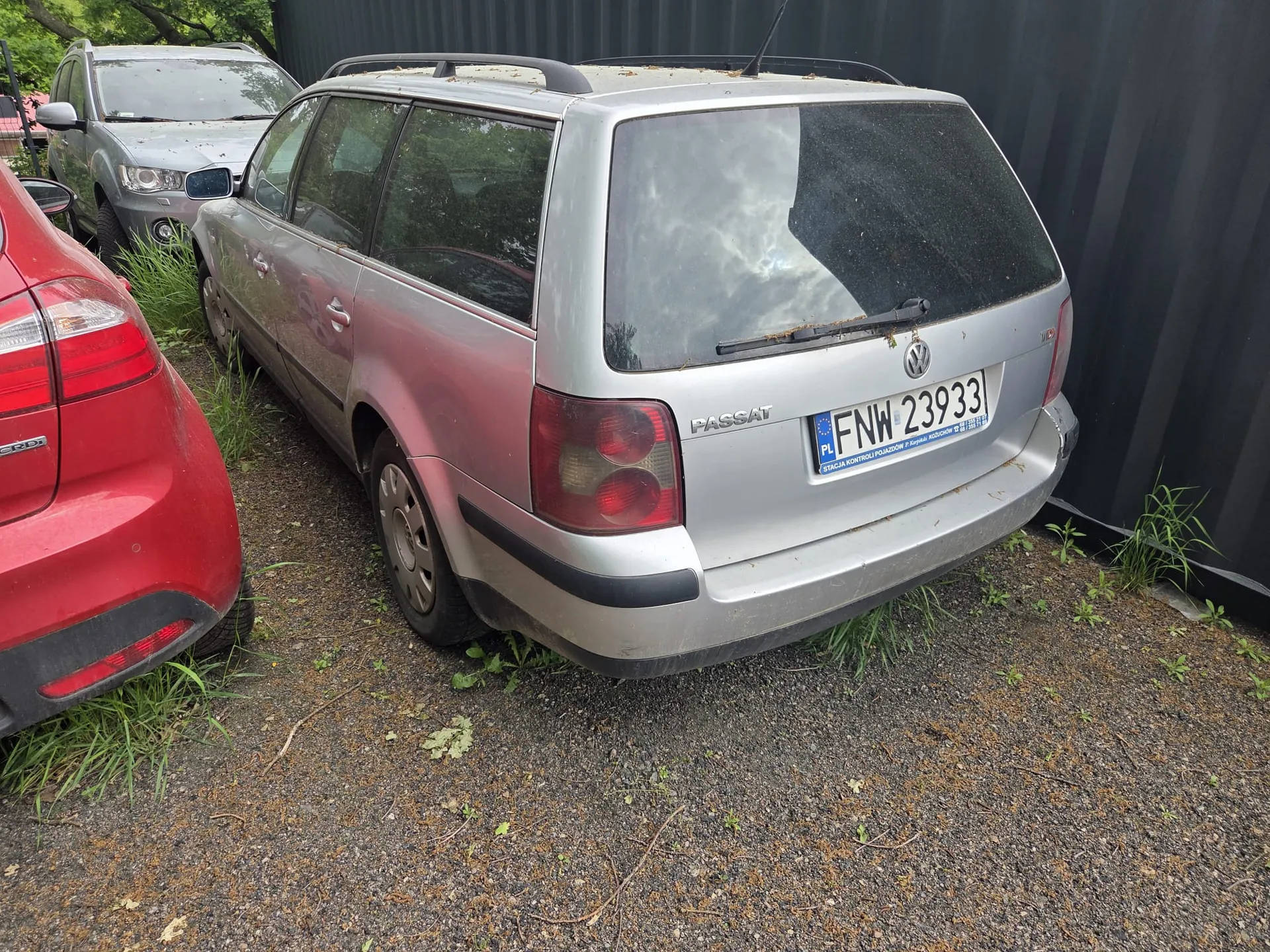 Srebrny samochód Volkswagen Passat kombi zaparkowany na żwirowym podłożu z trawą. Widoczny tył pojazdu z tablicą rejestracyjną PL FNW 23933 oraz napisem "PASSAT". Po lewej stronie czerwony samochód, w tle szary SUV. Tło stanowi ciemna metalowa bariera. Samochody są ustawione na nierównym terenie z rozproszoną trawą i liśćmi.