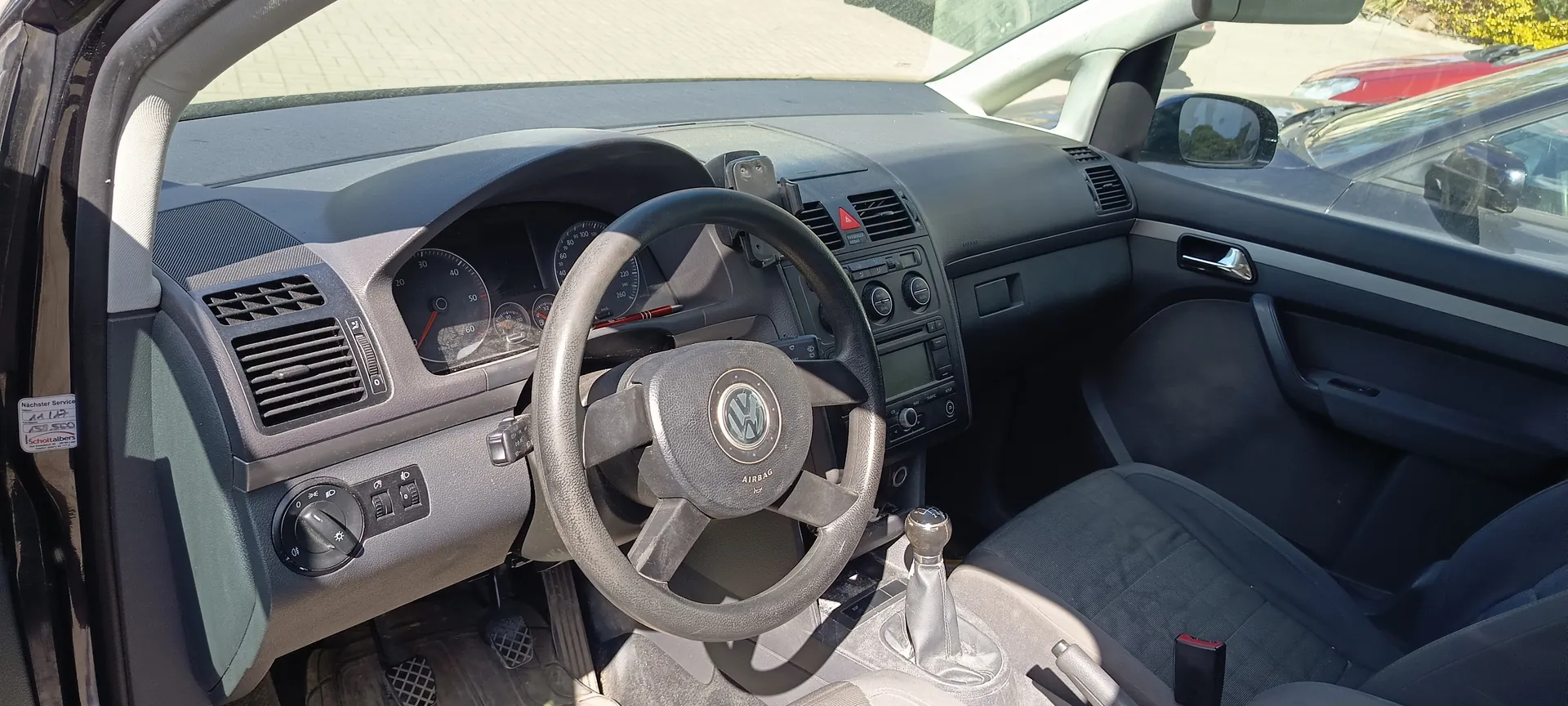 Wnętrze samochodu marki Volkswagen z widocznym kierownicą z logo VW i napisem AIRBAG. Deska rozdzielcza z licznikami prędkości i obrotomierzem, klimatyzacją oraz centralnym ekranem. Widoczny dźwignia zmiany biegów, siedzenia tapicerowane szarym materiałem. Na drzwiach pasażera boczne okno i uchwyt. Przez przednią szybę widoczne zaparkowane samochody oraz brukowana powierzchnia. Na drzwiach kierowcy naklejka z napisem Nächster Service 44 128 752 590 Scholten.