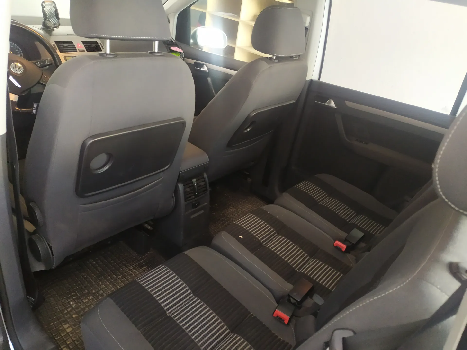 Wnętrze samochodu widziane z tyłu. Widoczne są dwa przednie siedzenia z podłokietnikami i zagłówkami oraz tylne siedzenia z paskami bezpieczeństwa o czerwonych zapięciach. Kierownica z logo Volkswagen po lewej stronie, deska rozdzielcza z przyciskami i wskaźnikami. Podłoga wyłożona matą z siateczkowym wzorem. Drzwi po prawej stronie z uchwytem i oknem. Wnętrze w odcieniach szarości i czerni.