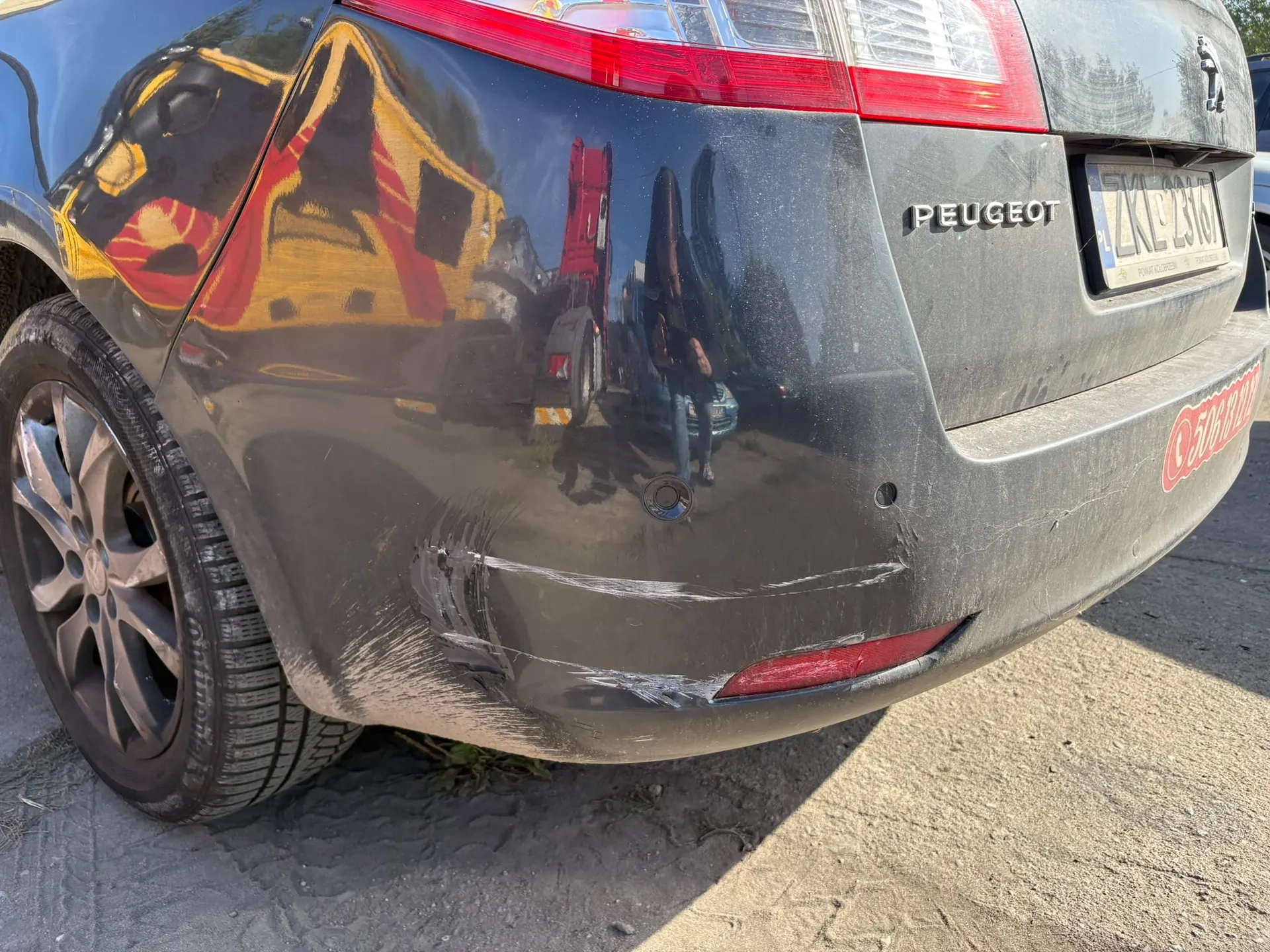 Zbliżenie na tylny lewy róg ciemnoszarego samochodu marki Peugeot. Na zderzaku widoczne są wyraźne uszkodzenia w postaci głębokich zarysowań i wgniecenia. W lakierze nadkola odbija się żółty pojazd serwisowy oraz postać osoby. Widoczny jest fragment tylnej lampy, napis PEUGEOT oraz fragment tablicy rejestracyjnej ZKL 23161.