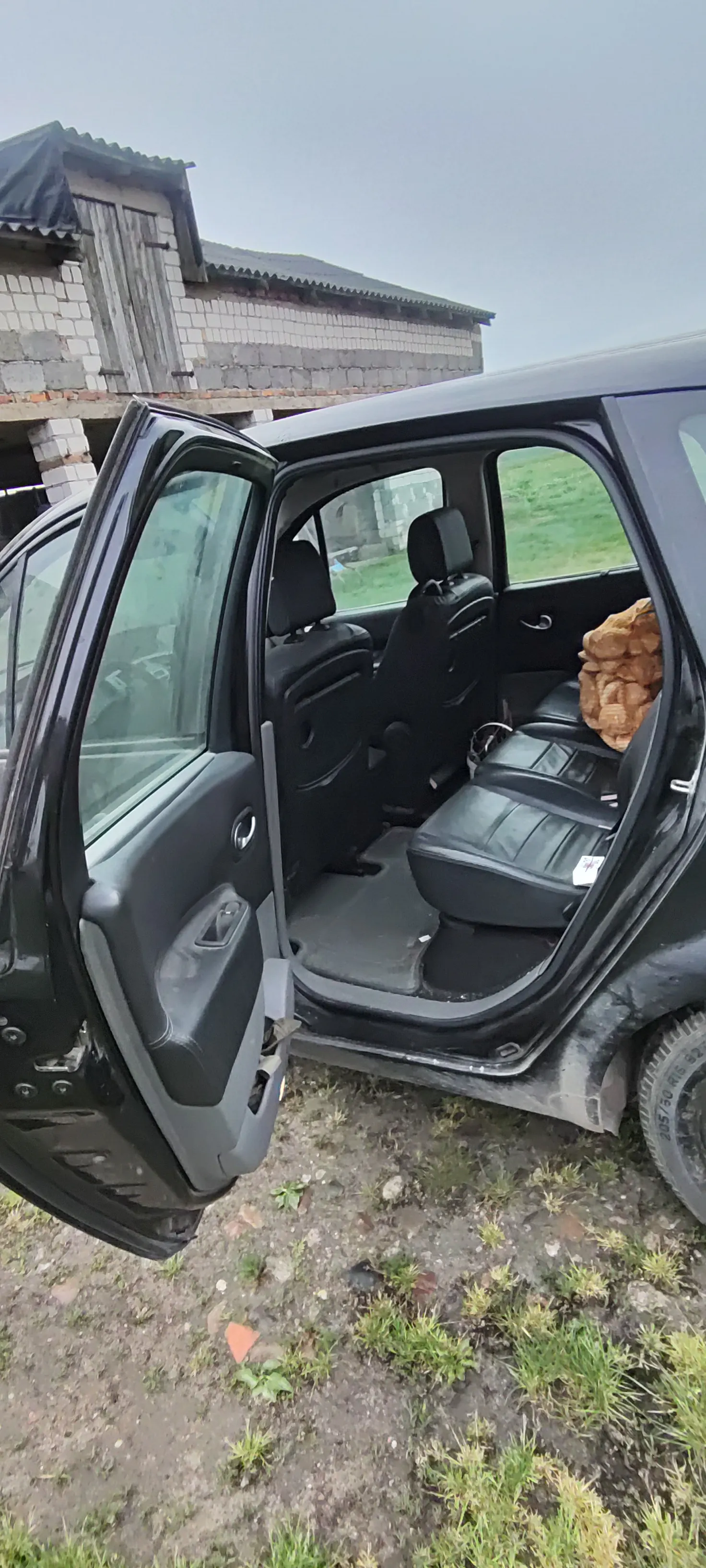Czarny samochód z otwartymi drzwiami, widoczne wnętrze z czarnymi siedzeniami. Na tylnym siedzeniu brązowy przedmiot. Samochód zaparkowany na brudnej ziemi z trawą. W tle budynek z cegły i szare niebo. Na oponie widoczny napis: 205/55 R16