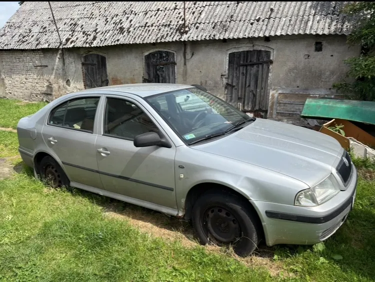 Srebrny samochód osobowy marki Volkswagen Bora zaparkowany na trawie przed starym, szarym budynkiem gospodarczym z trzema drewnianymi drzwiami i dachem z eternitu.