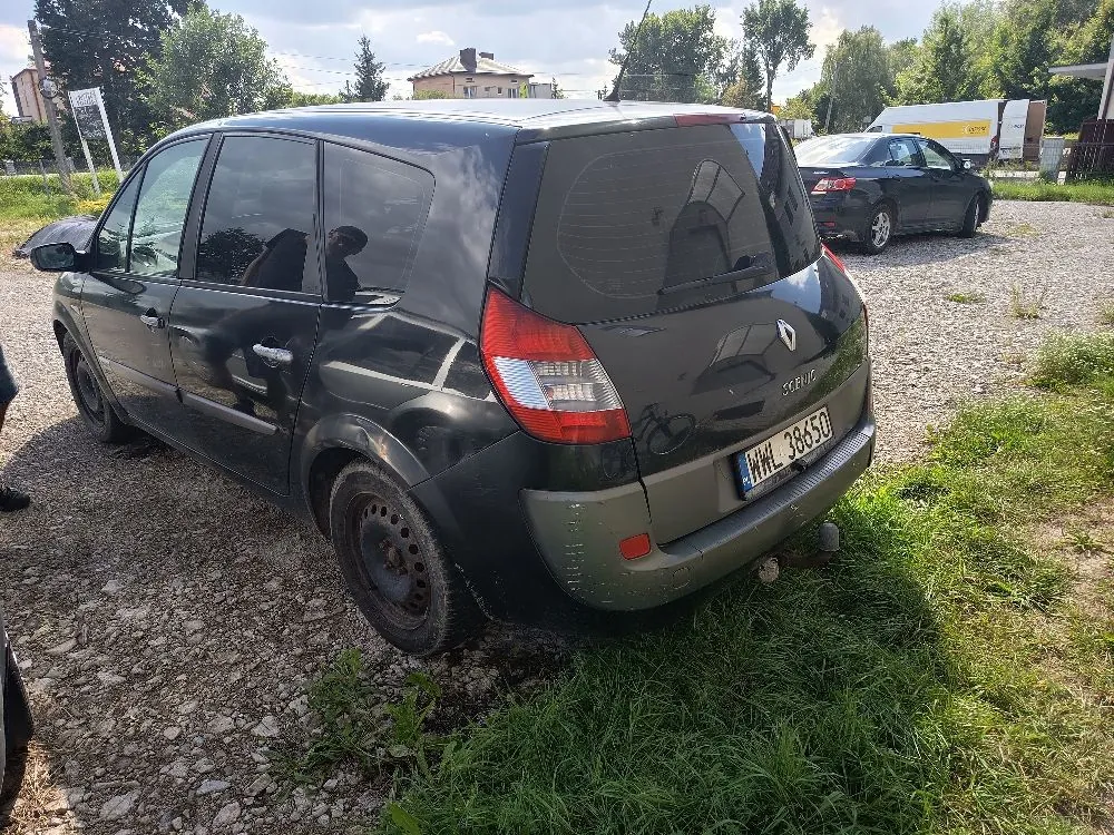 Ciemnozielony samochód marki Renault Scenic zaparkowany na żwirowym placu. Widoczny jest tył i lewy bok pojazdu. Samochód posiada polską tablicę rejestracyjną o numerze WWL 38650. W tle widać inny, ciemny samochód, zielone drzewa oraz fragmenty budynków pod jasnym niebem.