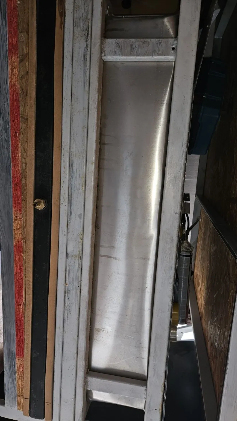 Metalowa konstrukcja z połyskującym stalowym panelem w centrum, otoczonym białymi metalowymi ramami. Nad panelem widoczne są elementy drewniane i narzędzia, w tym niebieskie pudełko oraz metalowy cylinder. Poniżej panelu znajdują się warstwy materiałów: biała pomalowana rama, deski drewniane, czarna taśma z mosiężnym elementem oraz czerwona farba na drewnie. Całość ma przemysłowy wygląd z widocznymi śladami zużycia i zabrudzeń.