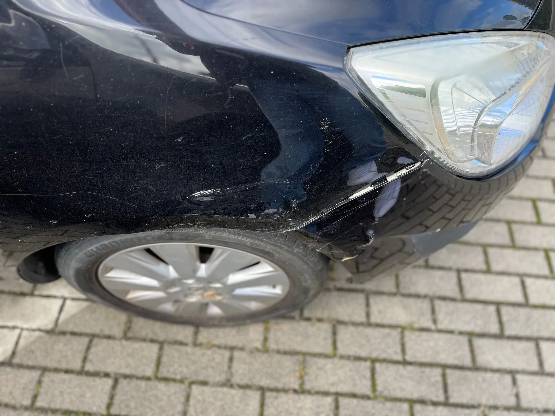 Zbliżenie na przedni prawy narożnik czarnego samochodu. Widoczne są zarysowania i wgniecenie na zderzaku w pobliżu reflektora oraz nad nadkolem. W kadrze znajduje się fragment przedniego koła z felgą aluminiową, samochód stoi na kostce brukowej.