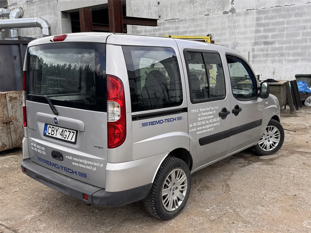 Srebrny samochód dostawczy marki Fiat Doblo zaparkowany na betonowym podłożu przed budynkiem z białych bloczków. Na tylnej klapie znajduje się tablica rejestracyjna o numerze CBY 4G77. Na boku i z tyłu pojazdu widnieją napisy firmowe: 'REMO-TECH' oraz dane kontaktowe: 'P.P.H.U. "Remo-Tech" sp. z o.o., ul. Przemysłowa 16, 69-010 Koronowo, tel. 052 3022 542, faks 052 3022 223, e-mail: remo-tech@remo-tech.pl, www.remo-tech.pl'.