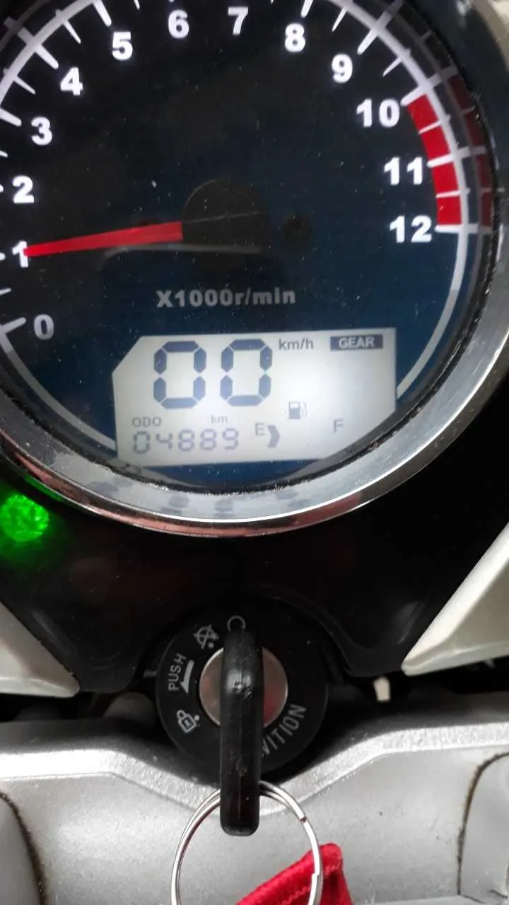 Zbliżenie na zegar samochodowy lub motocyklowy. Tachometr z zakresem od 0 do 12 (oznaczony jako X1000r/min), wskazówka na ok. 1000 obr./min. Cyfrowy wyświetlacz prędkości pokazuje 00 km/h. Licznik przebiegu (ODO) wskazuje 04889 km. Obok znajduje się wskaźnik paliwa z oznaczeniami E i F oraz napisem GEAR. Poniżej wyświetlacz, w stacyjce, włożony klucz z czerwoną etykietą i metalowym pierścieniem. Zielone światło jest włączone. Tekst i cyfry są czytelne, ustawione pionowo.