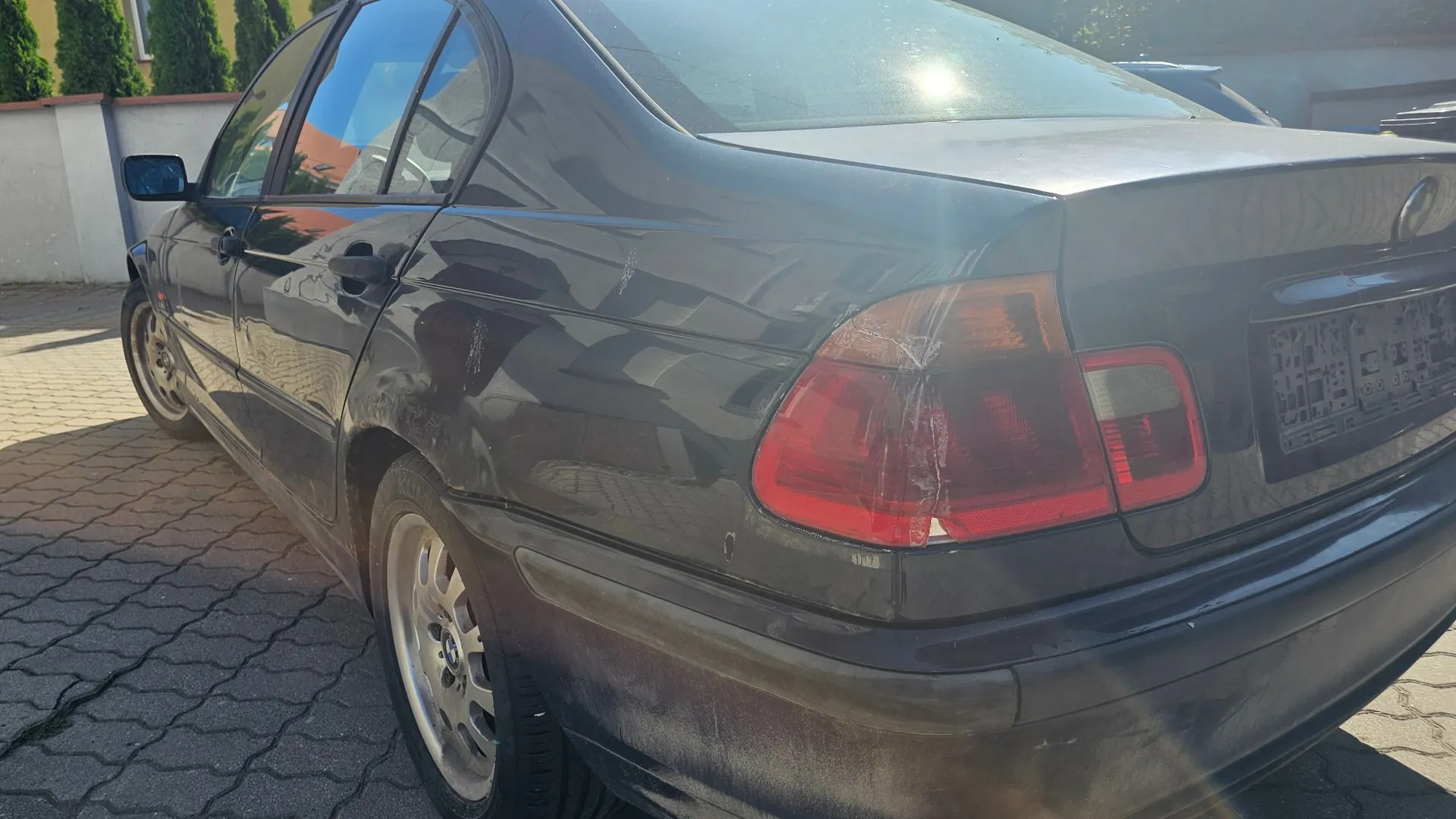 Tylna część i bok ciemnego samochodu osobowego marki BMW, zaparkowanego na kostce brukowej. Na nadwoziu widoczne są liczne zarysowania oraz uszkodzenia lakieru, szczególnie na tylnym błotniku i klapie bagażnika. Widoczna jest tylna lampa oraz tylne koło z oryginalną felgą.