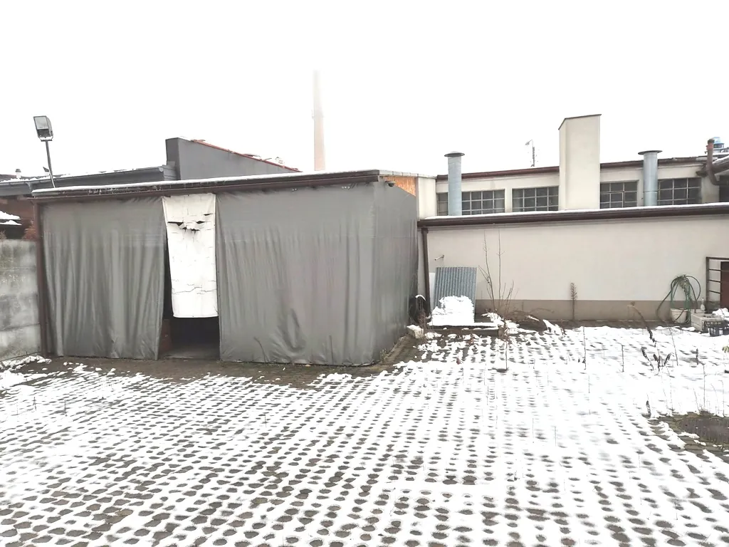 Zdjęcie przedstawia teren przemysłowy zimą, pokryty cienką warstwą śniegu. Na pierwszym planie znajduje się plac wyłożony betonową kratką trawnikową z białym śniegiem w zagłębieniach. Po lewej stronie stoi tymczasowa konstrukcja przypominająca magazyn lub garaż, osłonięta szarymi plandekami, z otwartym wejściem. W tle widoczny jest biały budynek przemysłowy z rzędami okien, kominami wentylacyjnymi oraz wysokim, cienkim kominem w oddali. Po prawej stronie budynku widać zwinięty zielony wąż ogrodowy.