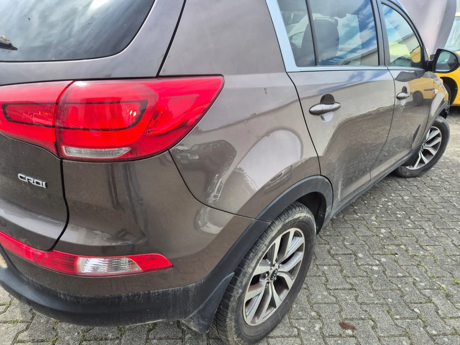 Zdjęcie tylnej, prawej części brązowego samochodu typu SUV. Widoczny jest tylny reflektor w kolorze czerwonym, napis 'CRDi' na klapie bagażnika oraz tylne prawe koło z aluminianą felgą. Samochód stoi na kostce brukowej.