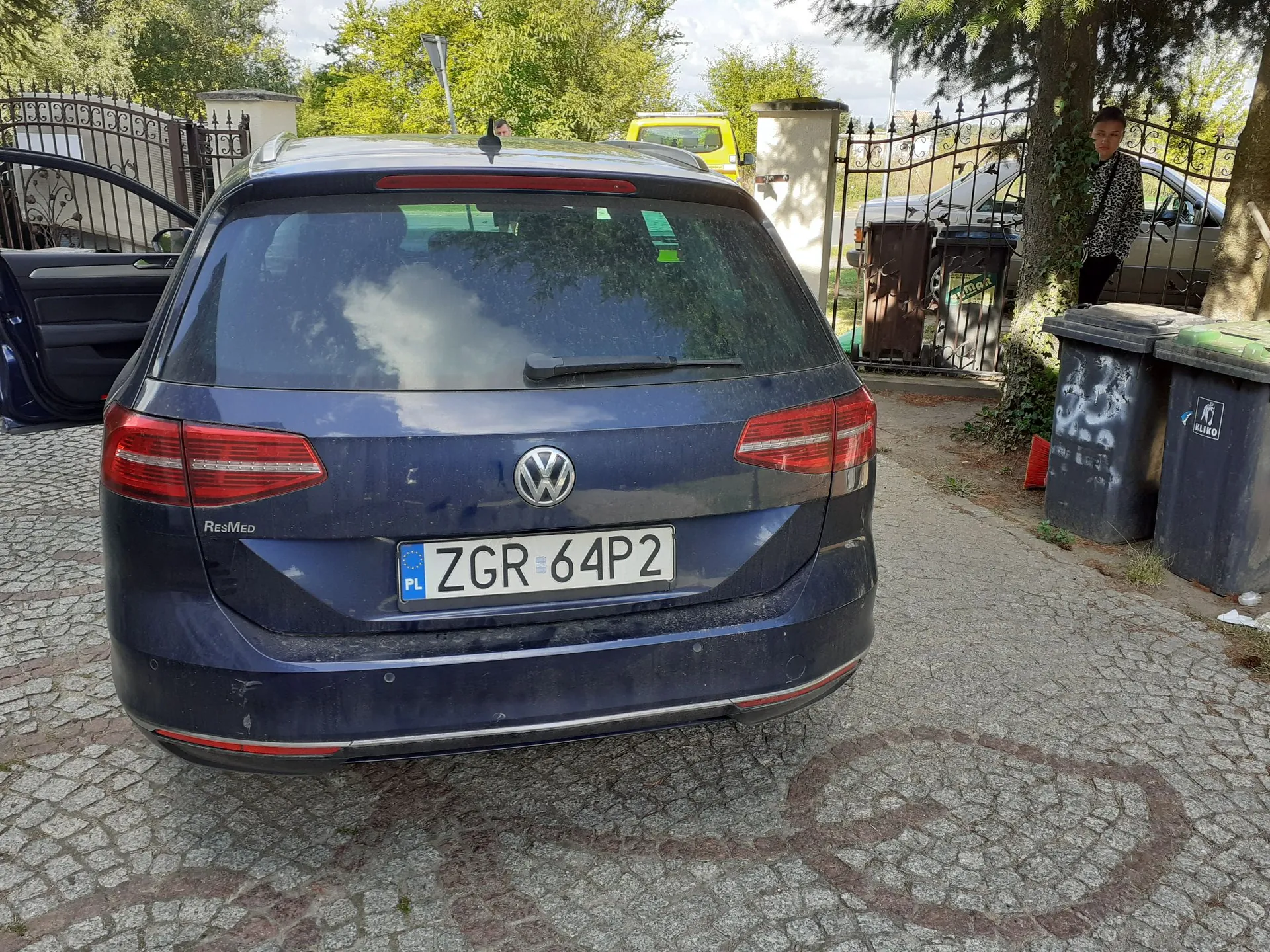Tylny widok ciemnoniebieskiego samochodu marki Volkswagen zaparkowanego na kostce brukowej. Samochód ma polską tablicę rejestracyjną o numerze ZGR 64P2. Po lewej stronie widoczny jest napis ResMed. W tle znajduje się metalowy płot, zielone drzewa, żółty samochód oraz osoba stojąca obok ciemnego samochodu i czarnych pojemników na odpady.