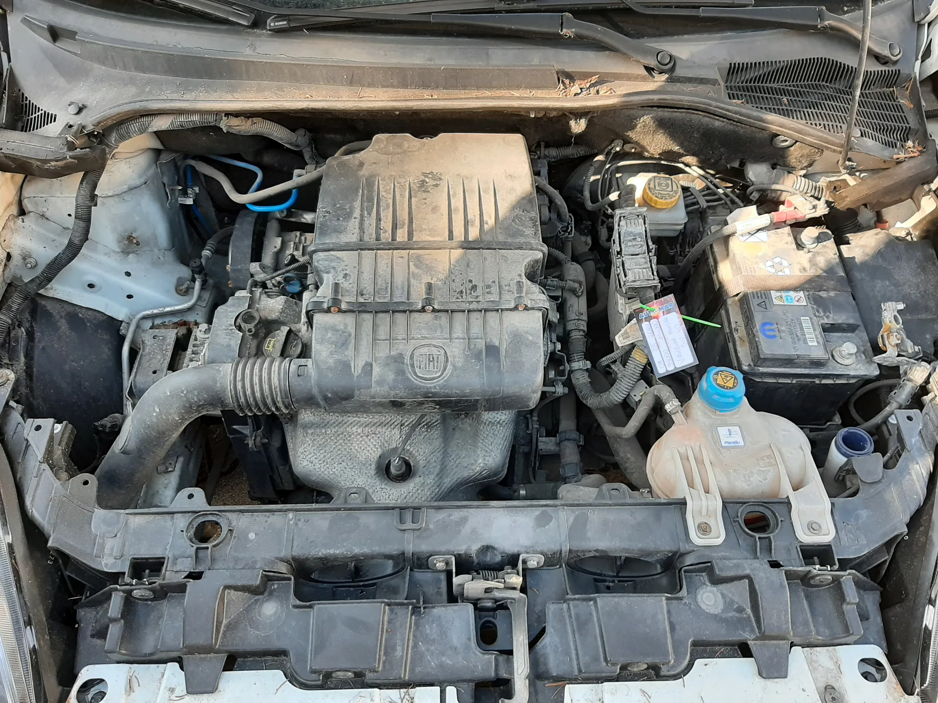 Widok z góry na zanieczyszczony silnik samochodu marki Fiat z otwartą maską. W centrum znajduje się pokrywa silnika z widocznym logo FIAT. Po prawej stronie widoczna jest bateria z oznaczeniami, kodem kreskowym oraz niebieską nakrętką zbiornika chłodniczego. Obok znajduje się żółta nakrętka wlewu oleju. Wokół rozłożone są przewody, węże i elementy mechaniczne. Na jednym z elementów umocowana jest biała etykieta z częściowo czytelnym napisem AUTO oraz zielonymi i czerwonymi znacznikami.