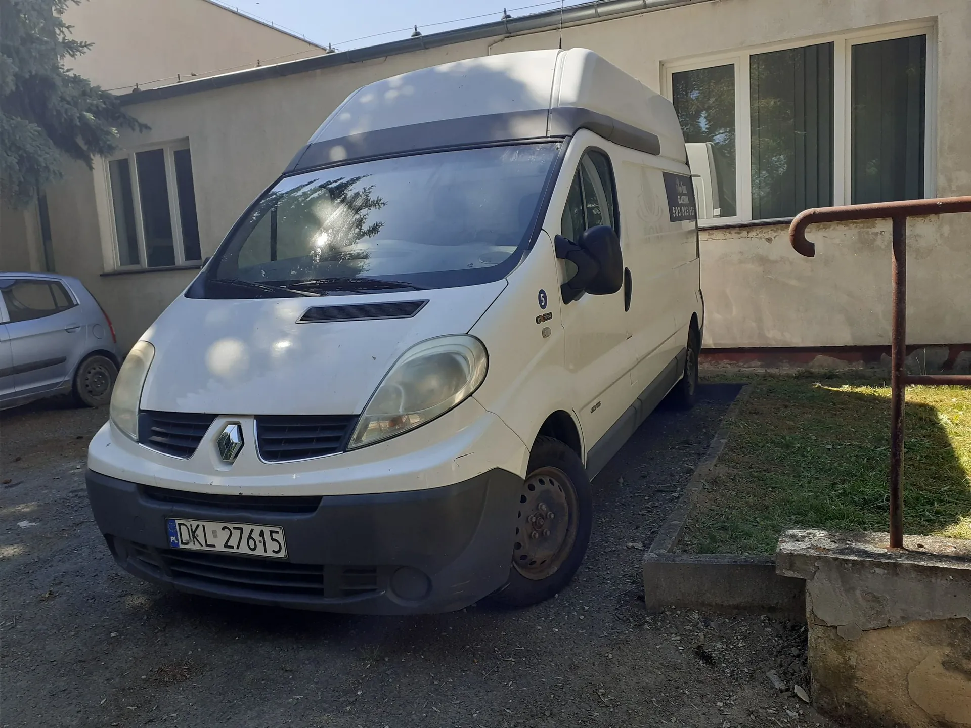 Biały furgon marki Renault zaparkowany przed jasnym budynkiem. Pojazd ma wysoką nadbudowę dachu i tablicę rejestracyjną o numerze DKL 27615. Po lewej stronie widoczny jest fragment innego, srebrnego samochodu, a po prawej metalowa barierka i fragment trawnika.