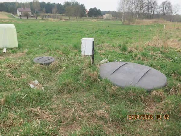 Zdjęcie przedstawiające teren zielony z trawą. Na pierwszym planie widoczne są dwie szare, owalne pokrywy włazów wbudowane w ziemię oraz biała, metalowa szafka elektryczna na słupku. W tle znajduje się otwarta przestrzeń, kilka budynków mieszkalnych oraz pas drzew. W prawym dolnym rogu widnieje data i godzina: 15/04/2026 10:02.