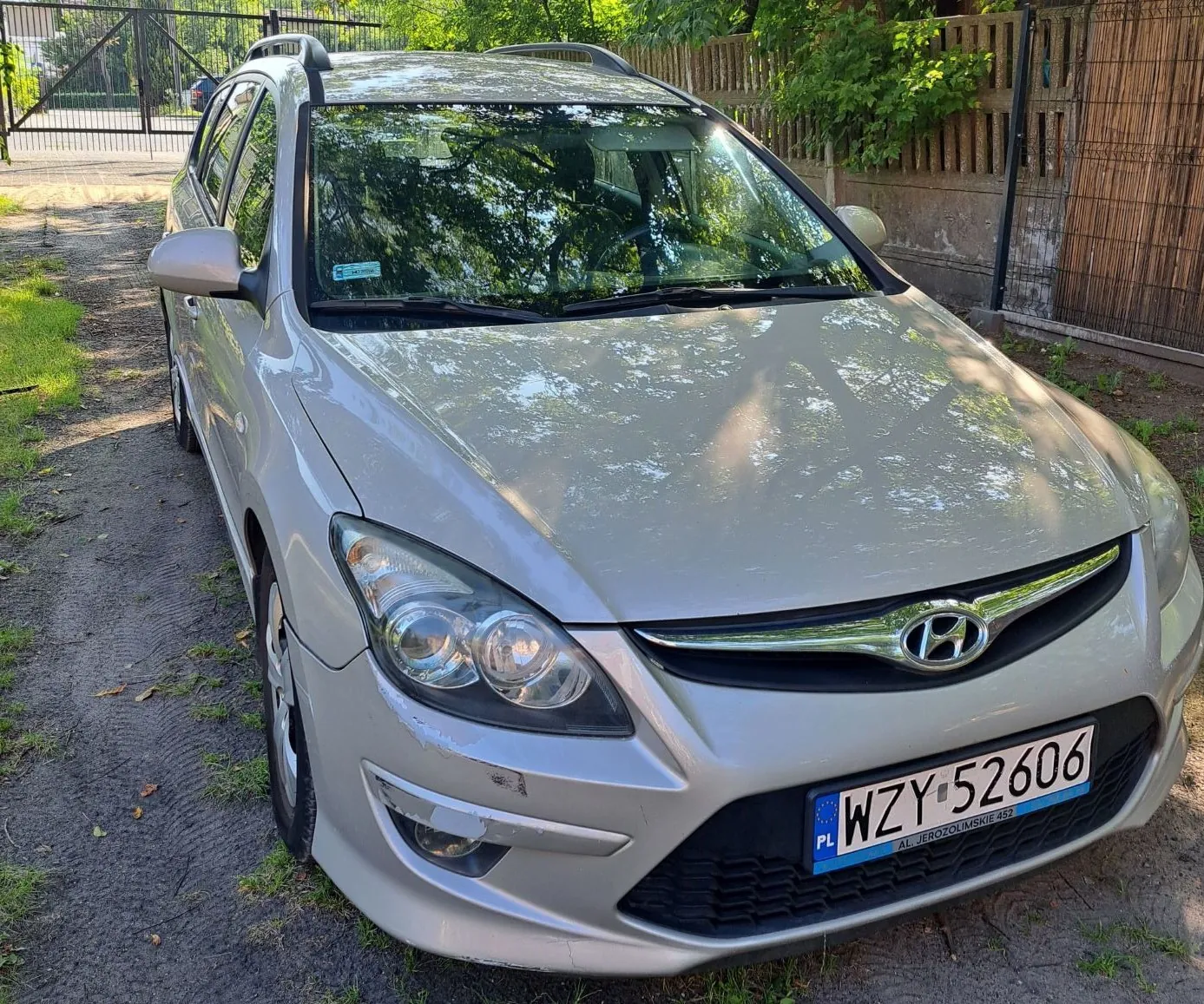 Srebrny samochód marki Hyundai w nadwoziu kombi, zaparkowany na piaszczystej drodze obok ogrodzenia. Widoczna przednia część pojazdu z tablicą rejestracyjną o numerze WZY 52606. Na masce i przedniej szybie odbijają się korony drzew.