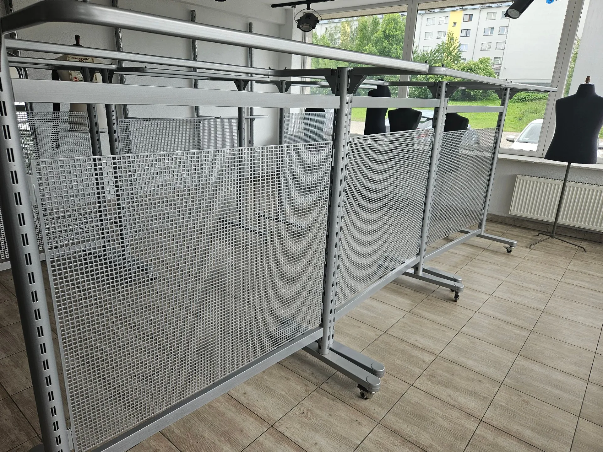 Srebrne, metalowe regały z siatkowymi ściankami ustawione w pomieszczeniu z jasną, kafelkową podłogą. W tle widoczne są duże okna z widokiem na budynki i zieleń oraz czarne manekiny krawieckie.