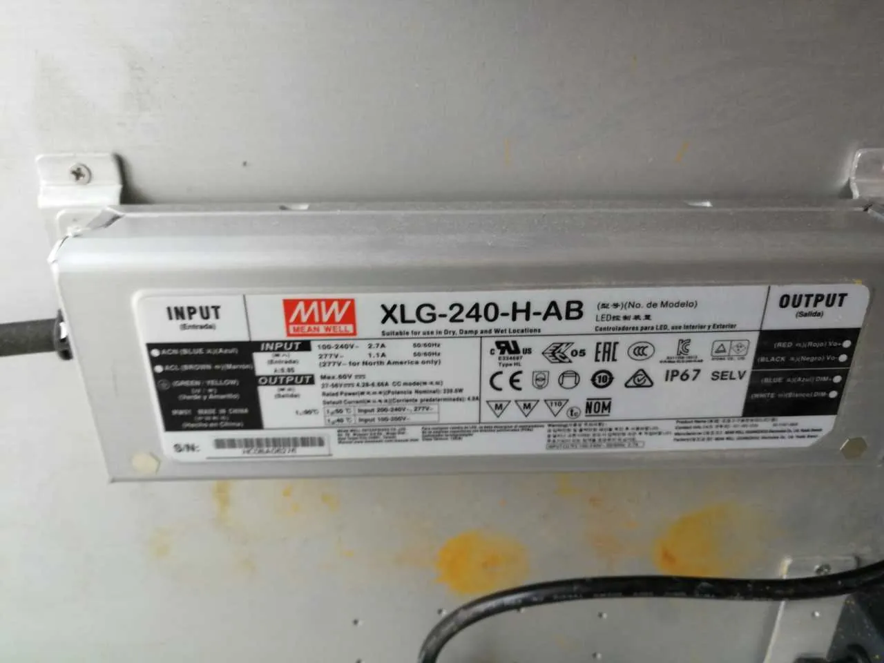Zdjęcie przedstawia metalowy element z białą etykietą zawierającą informacje techniczne. Na etykiecie widoczne są sekcje: INPUT (z kolorowymi oznaczeniami przewodów: niebieski, brązowy, zielono-żółty), logo MEAN WELL, model XLG-240-H-AB, opis przeznaczenia 'LED control device', parametry wejściowe (100-240V~, 2.7A) i wyjściowe (27-56V, 4.28-8.64A), oznaczenia certyfikatów (CE, EAC, IP67, SELV) oraz dodatkowe symbole bezpieczeństwa. W dolnej części etykiety znajduje się numer seryjny (S/N) i kod kreskowy. Element jest zamocowany na powierzchni z widocznymi śrubami i przewodami po bokach. Tło stanowi metalowa powierzchnia z plamami.
