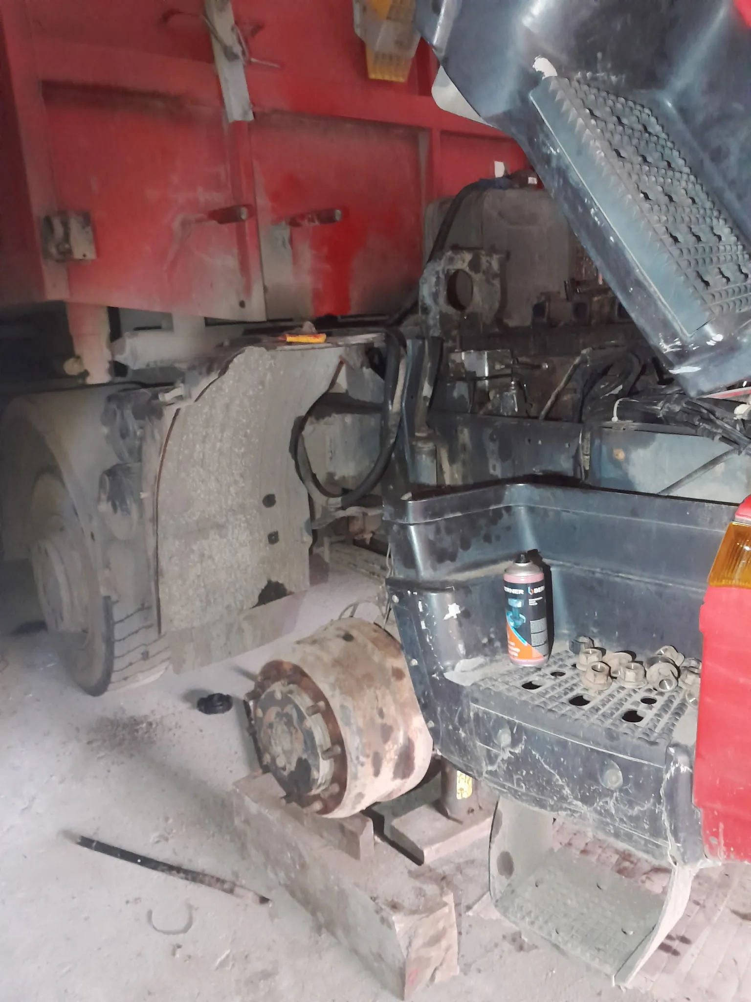 Czerwony ciężarówka z otwartą maską silnika, widoczne elementy mechaniczne, w tym zardzewiały felga, butelka sprayu z częściowo czytelnym napisem 'BRAK' oraz narzędzia na betonowej podłodze. W tle czerwona kabina pojazdu i elementy warsztatowe.