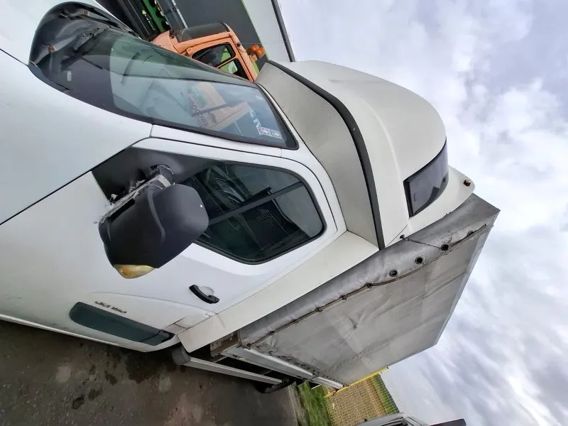 Biały samochód dostawczy z przyczepą o pokrytej tkaniną ładowni, zaparkowany na nawierzchni asfaltowej. Widoczne uszkodzenie lusterka bocznego. Na przedniej szybie napis '512'. W tle pomarańczowy pojazd, ogrodzenie i zachmurzone niebo.