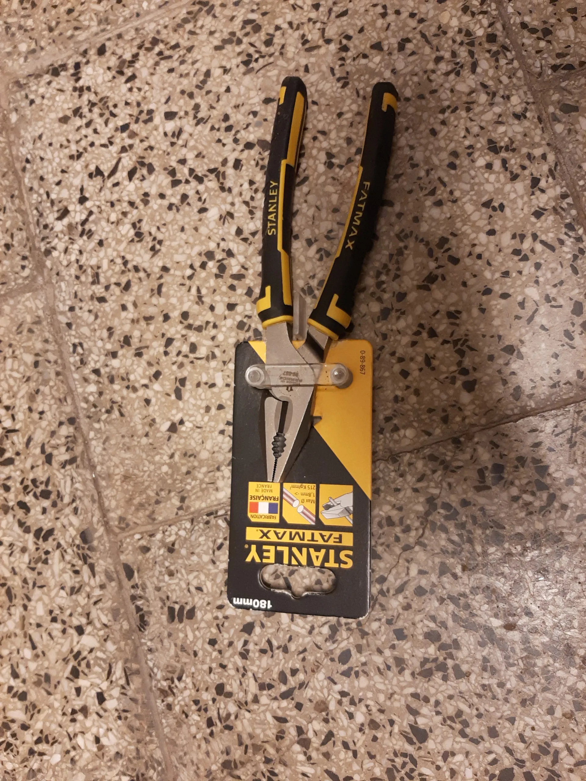 Parę szczypiec marki Stanley FatMax o długości 180 mm w opakowaniu. Szczypce mają czarno-żółte uchwyty z napisem 'STANLEY' i 'FATMAX'. Opakowanie jest czarno-żółte z widocznym tekstem 'STANLEY FATMAX', '180mm', 'FABRICATION FRANCAISE' oraz flagą Francji. Na opakowaniu znajduje się również numer '0-89-867' i schematyczne ilustracje zastosowań. Tło stanowi posadzka z kruszywem w kolorach beżowym i czarnym.