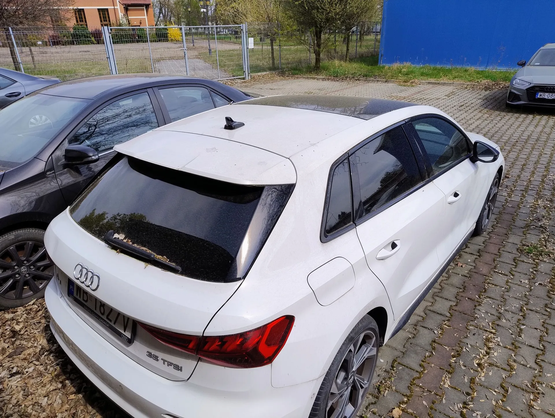 Biały samochód marki Audi w nadwoziu hatchback zaparkowany na kostce brukowej. Widoczny jest tył i prawy bok pojazdu. Na klapie bagażnika znajduje się logo Audi, tablica rejestracyjna o numerze ND 1827V oraz oznaczenie modelu 35 TFSI. Obok stoi ciemnoszary samochód. W tle widać ogrodzenie z siatki, drzewa oraz fragment niebieskiego budynku.