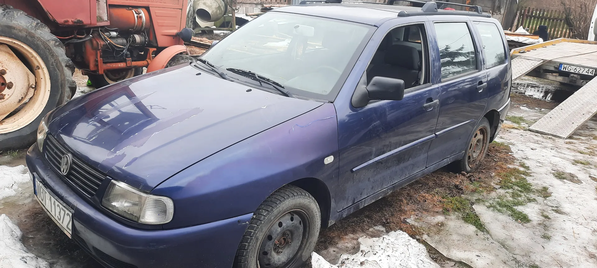Niebieski samochód osobowy typu kombi marki Volkswagen z tablicą rejestracyjną WOT 11372, zaparkowany na podjeździe z resztkami śniegu i trawą. Po lewej stronie widoczna część czerwonego ciągnika lub maszyny rolniczej z dużym kołem. W tle po prawej stronie przyczepa z tablicą rejestracyjną WG-62744. Samochód ma zarysowane uszkodzenia na karoserii i brudne felgi. Tło obejmuje ogrodzenie i fragmenty innych obiektów.