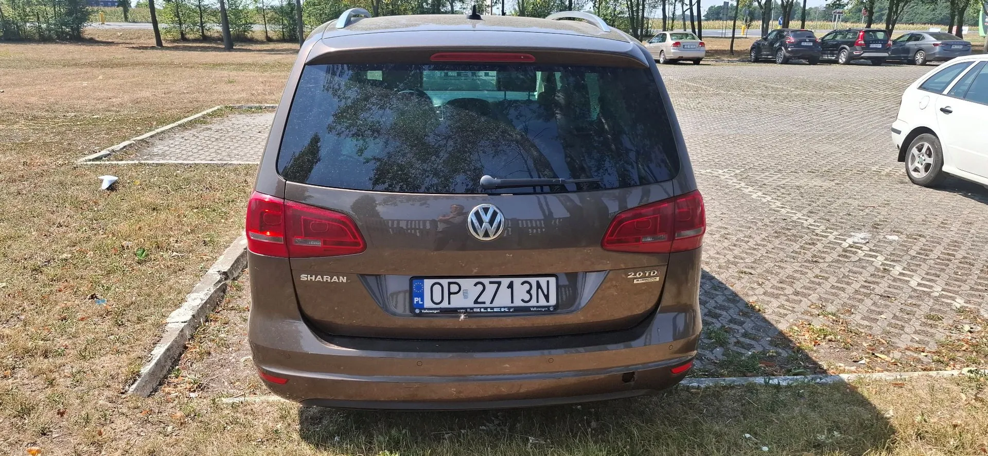 Tylny widok brązowego samochodu marki Volkswagen Sharan zaparkowanego na placu. Na klapie bagażnika znajduje się logo Volkswagena, napis 'SHARAN' po lewej stronie oraz '2.0 TDI' po prawej. Samochód ma polską tablicę rejestracyjną o numerze OP 2713N. W tle widoczne są inne zaparkowane samochody oraz pas zieleni z drzewami.