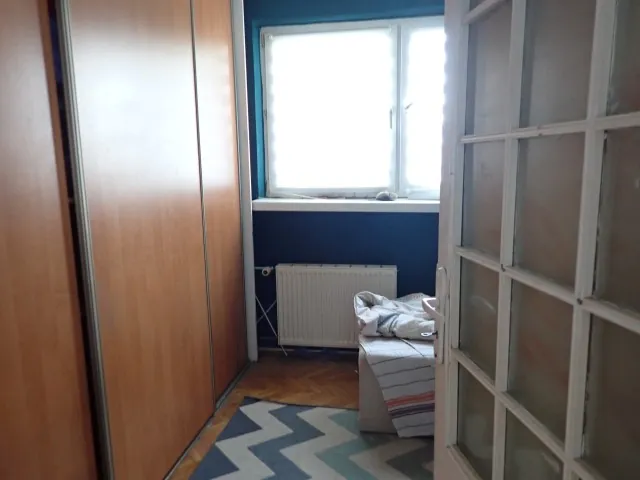 Wnętrze pokoju z widoczną drewnianą szafą przesuwną po lewej stronie, białym grzejnikiem pod oknem oraz fragmentem łóżka z pościelą. Na podłodze z parkietu leży dywan w geometryczne wzory w odcieniach niebieskiego, szarego i białego. Po prawej stronie znajduje się białe drzwi z przeszkleniami w kratkę.
