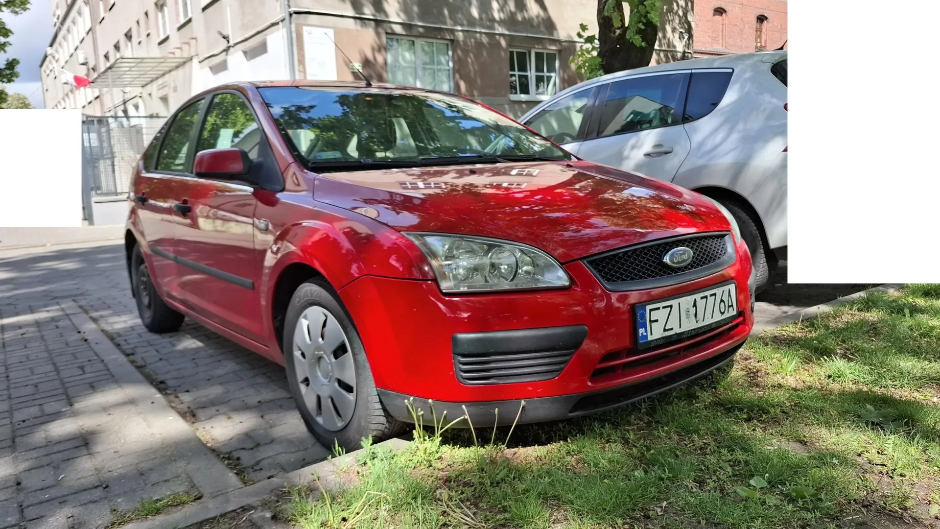 Czerwony samochód marki Ford zaparkowany na trawniku obok chodnika. Pojazd posiada polską tablicę rejestracyjną o numerze FZI 1776A. W tle widoczny jest biały samochód oraz jasna elewacja budynku z oknami.