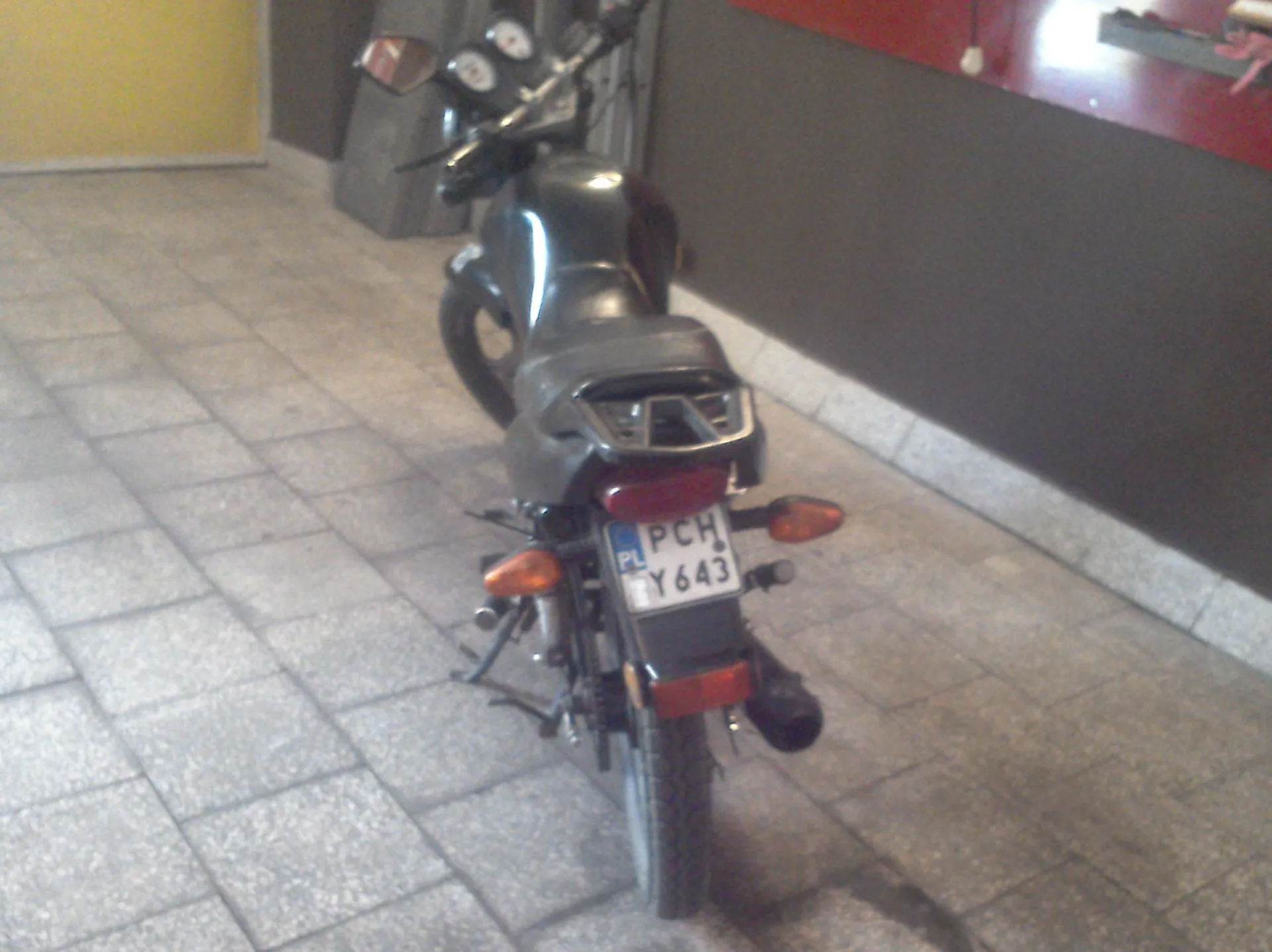 Widok z tyłu na czarny motocykl zaparkowany na szarym, kostkowym chodniku. Motocykl posiada polską tablicę rejestracyjną o numerze PCH Y643. W tle widoczna jest ciemna ściana oraz fragment żółtej ściany po lewej stronie.