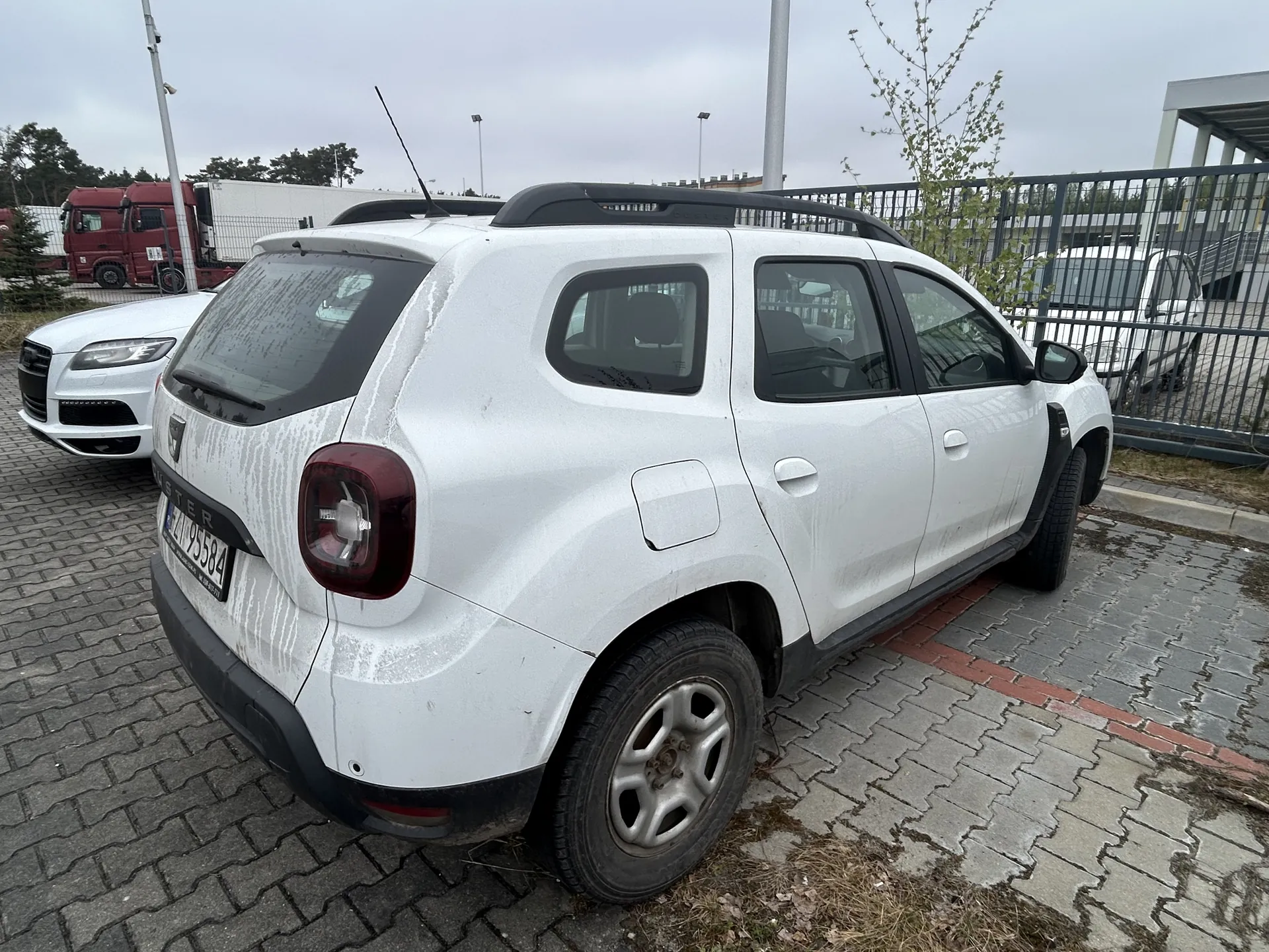 Biały samochód typu SUV zaparkowany na brukowanym placu. Widoczne są zabrudzenia na nadwoziu, czerwone światło tylnego reflektora oraz tablica rejestracyjna ZM 95584. W tle drugi biały samochód, czerwona ciężarówka, metalowe ogrodzenie, budynki przemysłowe i drzewa. Niebo jest zachmurzone.