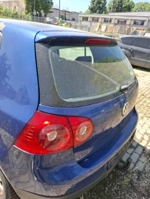 Tylna część niebieskiego samochodu osobowego. Widoczna jest tylna szyba, prawe światło stopu oraz fragment zderzaka z hakiem holowniczym. W tle znajdują się budynki i drzewa.