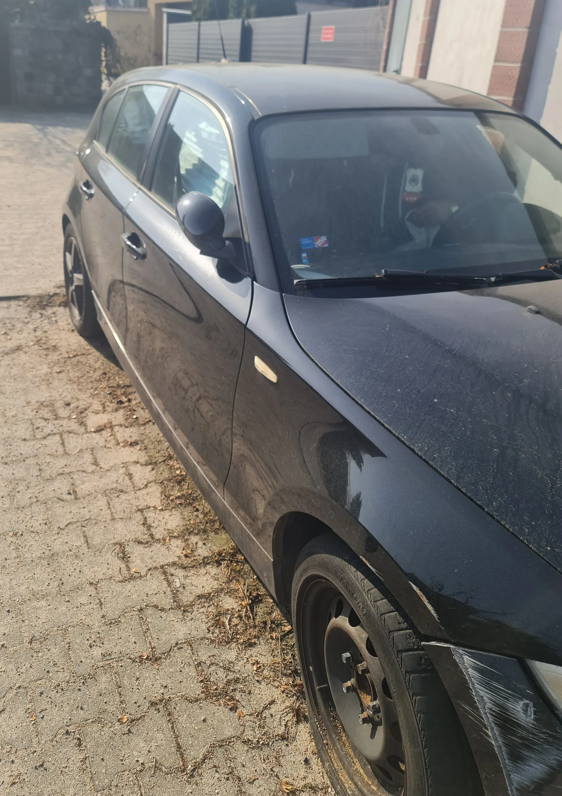 Czarny samochód osobowy typu hatchback zaparkowany na brukowanej powierzchni z suchymi liśćmi. Widoczne uszkodzenie przedniego zderzaka owinięte plastikową folią. Na przedniej szybie naklejki, w tym niebieska i czerwona. W tle budynki z ceglaną ścianą i metalowym ogrodzeniem. Opona przedniego koła z widocznymi śladami brudu i rdzy na felgach.