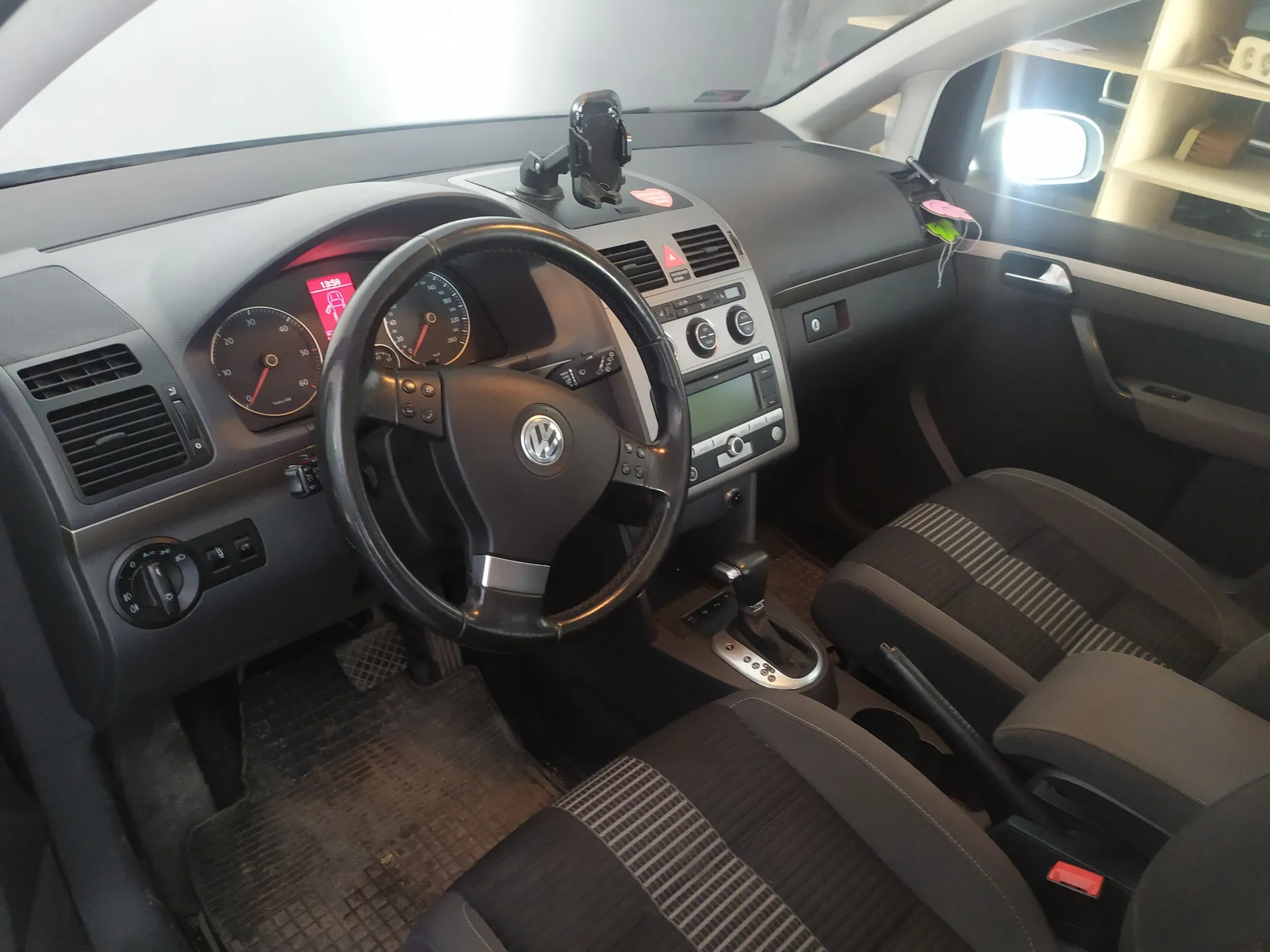 Wnętrze samochodu marki Volkswagen z widoczną kierownicą z logo VW, zegarami samochodowymi (w tym prędkościomierzem i tachometrem z czerwonymi wskazówkami), centralną konsolą z ekranem, klimatyzacją i przyciskami. Na desce rozdzielczej znajduje się uchwyt na telefon oraz naklejki. Widoczne są czarne siedzenia z paskowanym wzorem, dźwignia zmiany biegów i brudne dywaniki. Na wyświetlaczu deski rozdzielczej widnieje czas 13:58 oraz ikona samochodu. W tle przez przednią szybę widać pomieszczenie z półkami.