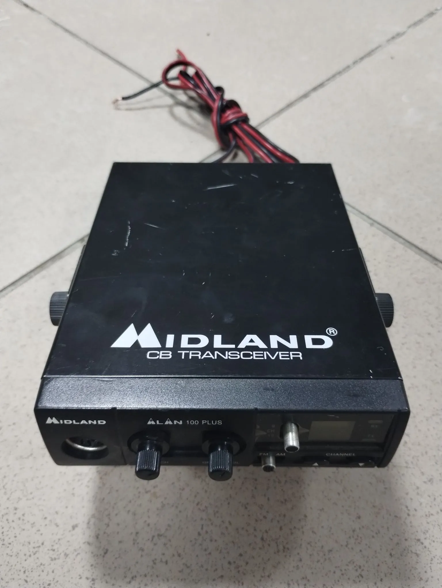 Czarny transceiver CB marki Midland, model Alan 100 Plus. Urządzenie leży na jasnej, kafelkowej powierzchni. Na górnej obudowie znajduje się biały napis 'MIDLAND CB TRANSCEIVER'. Panel przedni zawiera logo 'MIDLAND', nazwę modelu 'ALAN 100 PLUS', dwa pokrętła regulacji, gniazdo mikrofonu oraz gniazdo antenowe. Z tyłu urządzenia widoczne są czerwone i czarne przewody zasilające.